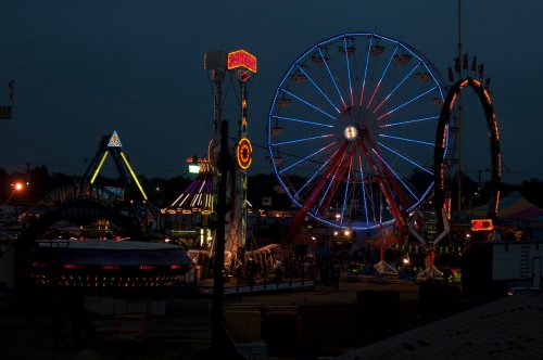 A vibrant carnival scene at dusk, featuring a large illuminated Ferris wheel and colorful rides.