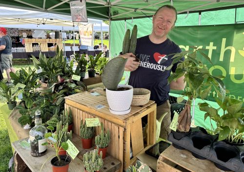 A smiling vendor stands behind a table filled with various potted plants at a market under a green tent.