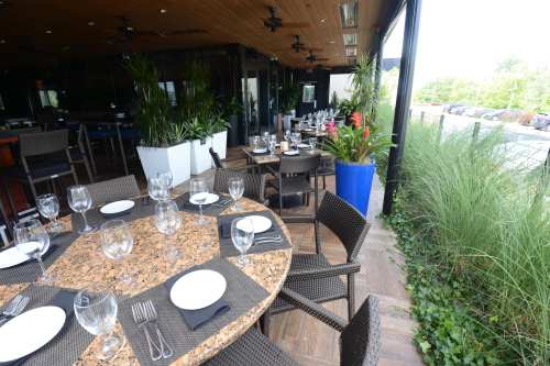 Outdoor dining area with a round table set for guests, surrounded by greenery and a modern restaurant ambiance.