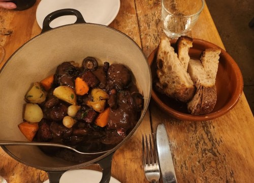 A hearty stew with vegetables in a pot, served with slices of bread on the side, on a rustic wooden table.