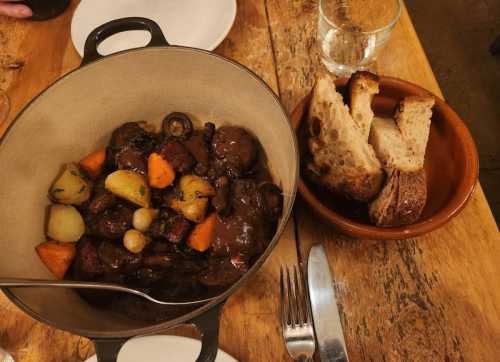 A hearty stew with vegetables in a pot, served with slices of bread on the side, on a rustic wooden table.