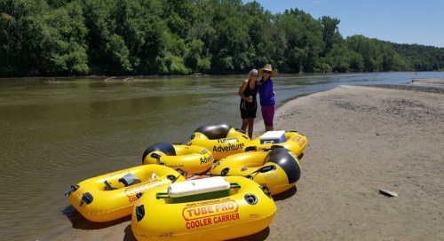 Two people stand on a sandy riverbank beside yellow inflatable tubes, with trees lining the river in the background.