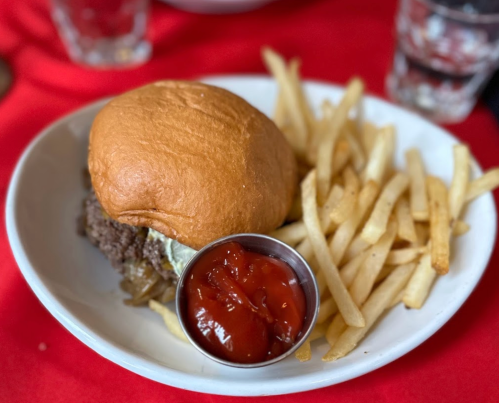 A plate with a burger topped with pickles and a side of golden fries, accompanied by a small bowl of ketchup.