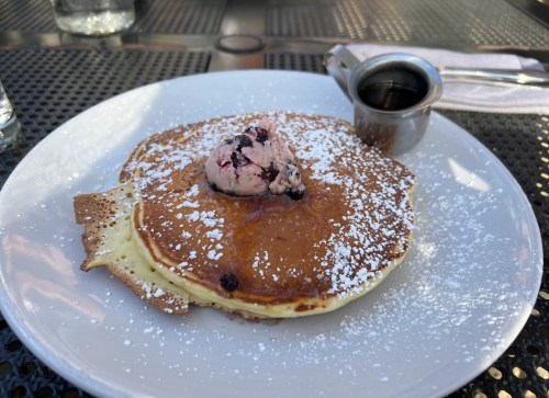 A plate with a fluffy pancake topped with a scoop of berry ice cream and a drizzle of syrup, dusted with powdered sugar.