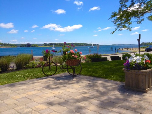A scenic view of a waterfront with flowers in a bicycle planter, blue sky, and boats in the distance.