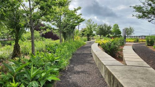 A winding path through a lush green park with trees and plants under a cloudy sky.