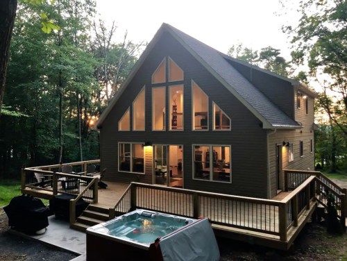 A modern cabin with large windows, a deck, and a hot tub, surrounded by trees at sunset.