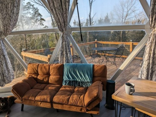 Cozy interior of a geodesic dome with a brown couch, blue throw, and a view of a wooden deck and trees outside.