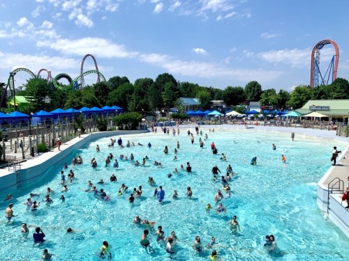 A busy water park with a large pool filled with people, surrounded by trees and amusement rides in the background.