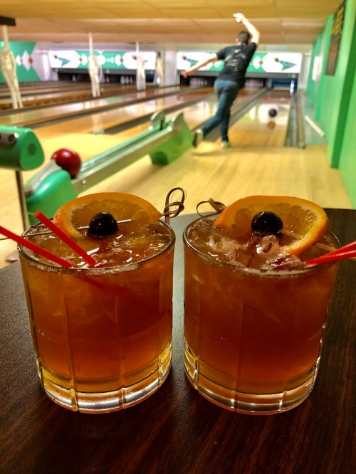 Two cocktails with orange slices and cherries sit on a table, with a bowler in the background at a bowling alley.