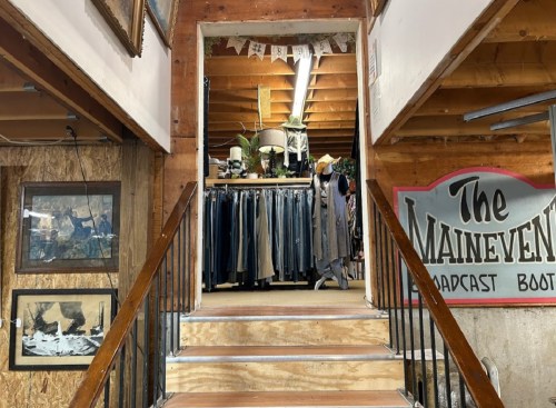 A staircase leads to a clothing store with racks of clothes and vintage decor in a rustic setting.