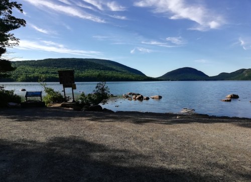 A serene lake surrounded by green hills under a clear blue sky, with rocks and a sign near the water's edge.