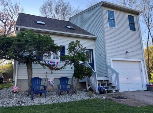 A two-story house with a garage, blue chairs in front, and a landscaped yard featuring a small tree and flowers.