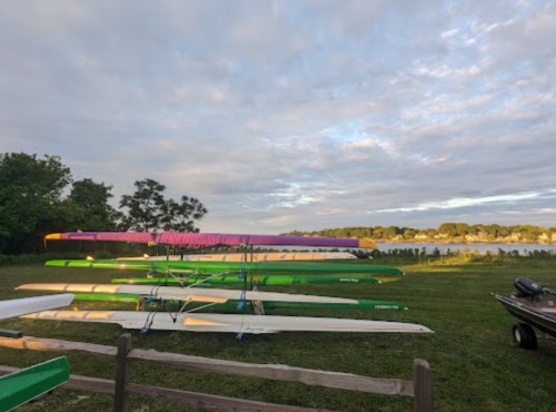 Rowing boats stacked on a grassy area by a calm lake under a cloudy sky.