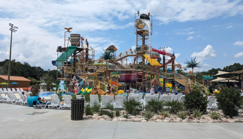 A colorful water park with slides and play structures, surrounded by lounge chairs and palm trees under a cloudy sky.