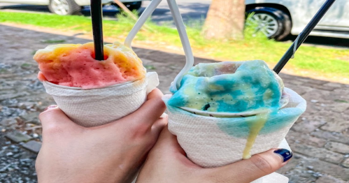 Two hands holding cups of colorful ice cream, one pink and orange, the other blue and yellow, wrapped in napkins.