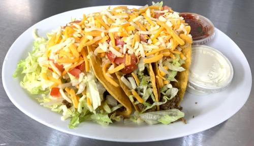 Three tacos filled with lettuce, tomatoes, and cheese, served with salsa and sour cream on a white plate.