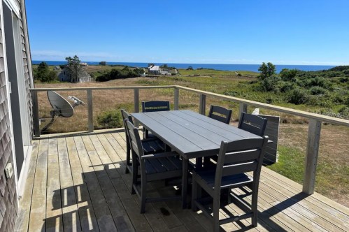 A wooden deck with a gray dining table and chairs, overlooking a grassy landscape and the ocean in the distance.