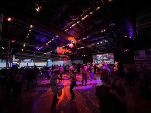 A lively dance floor with people dancing under colorful lights and a disco ball in a large venue.