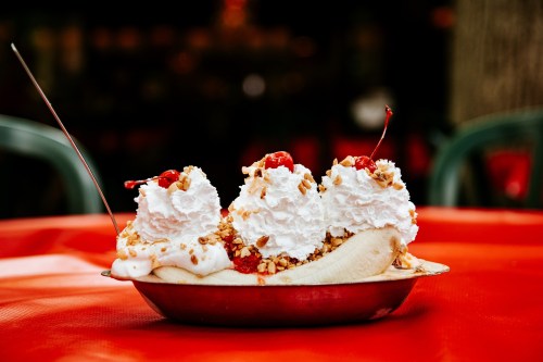 A banana split topped with whipped cream, cherries, and nuts, served in a metal dish on a red tablecloth.