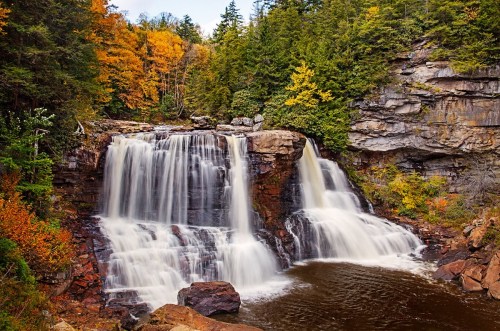 A serene waterfall cascades over rocks, surrounded by vibrant autumn foliage and a calm pool below.