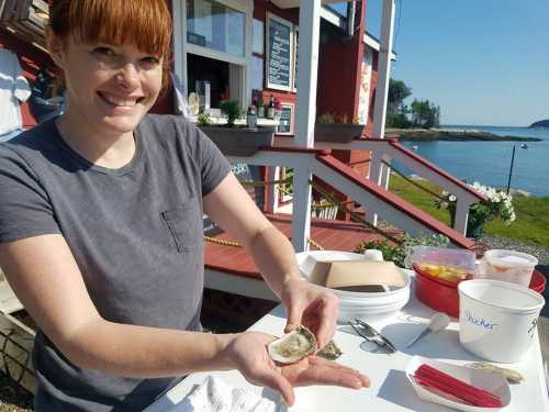 A smiling woman holds an open oyster at a seaside restaurant, with a scenic view in the background.