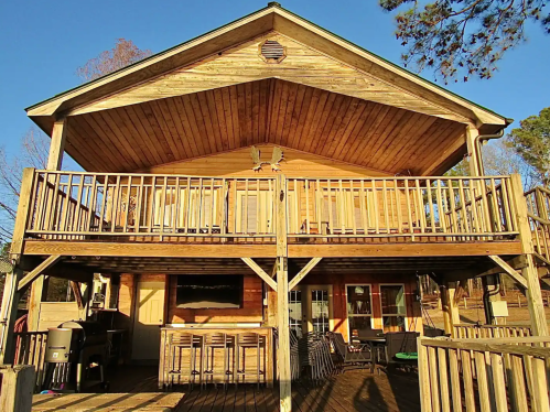 A wooden two-story house with a large deck and railing, surrounded by trees under a clear blue sky.
