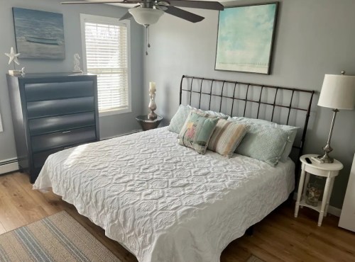 Cozy bedroom featuring a bed with a white quilt, dresser, lamp, and artwork on the walls. Natural light from windows.