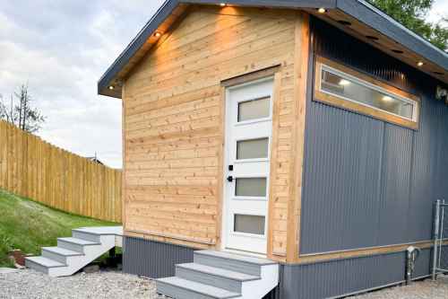 A modern tiny house with a wooden and metal exterior, featuring a white door and steps leading up to it.