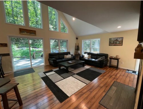 Spacious living room with large windows, black sofas, a coffee table, and wooden floors, overlooking a green outdoor area.