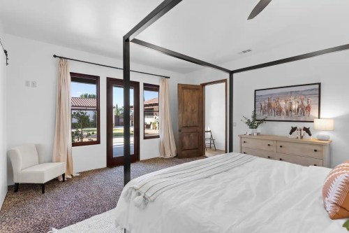 Bright bedroom with a four-poster bed, large windows, and a neutral color palette, featuring a cozy chair and artwork.