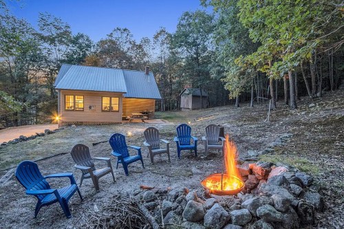A cozy cabin surrounded by trees, with a fire pit and blue chairs arranged in a circle, set in a serene outdoor setting.