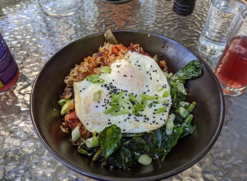 A bowl of rice topped with two fried eggs, sautéed greens, and garnished with green onions and black sesame seeds.
