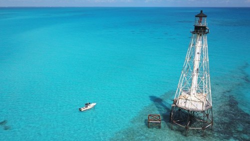 A white lighthouse stands on a platform in vibrant turquoise waters, with a small boat nearby under a clear blue sky.