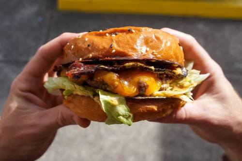 A person holds a large burger with lettuce, cheese, and crispy bacon, featuring a sesame seed bun.