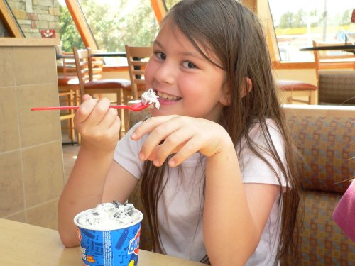 A smiling girl with long hair enjoys a cup of ice cream, holding a spoon and sitting in a bright café.