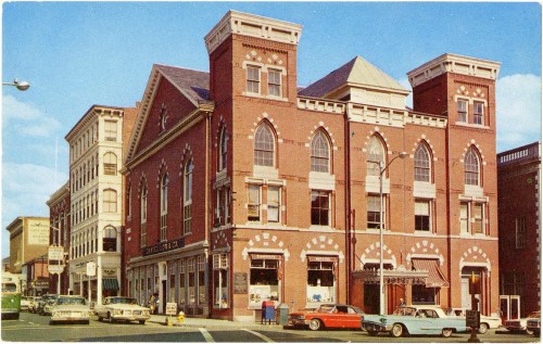 Historic brick building with arched windows, surrounded by vintage cars and shops on a sunny day.