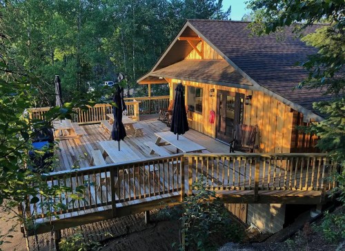A wooden cabin with a spacious deck surrounded by trees, featuring picnic tables and umbrellas.