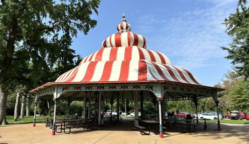 A striped red and white pavilion with a domed roof, surrounded by trees and picnic tables on a sunny day.