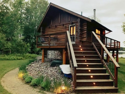 A cozy log cabin with a deck, surrounded by greenery and a stone pathway, illuminated by soft outdoor lights.