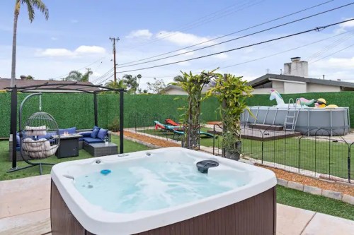 A backyard scene featuring a hot tub, lounge area, and a pool with colorful floats, surrounded by greenery.
