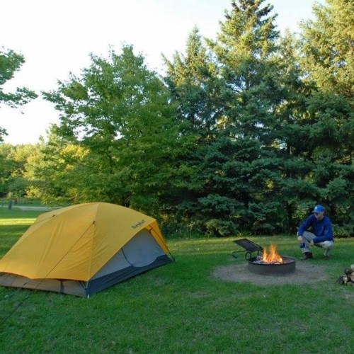 A yellow tent beside a campfire in a green park, surrounded by trees and grass. A person sits near the fire.