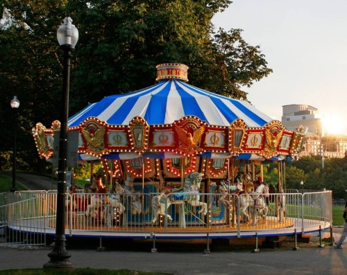 A colorful carousel with ornate horses, surrounded by a fence, set against a sunset in a park.