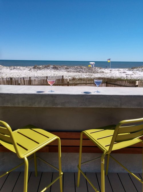 Two green bar stools overlook a sandy beach and ocean, with two cocktails on a ledge under a clear blue sky.