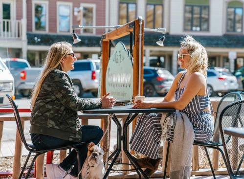 Two women sit at an outdoor café table, chatting and enjoying drinks, with a dog resting nearby.