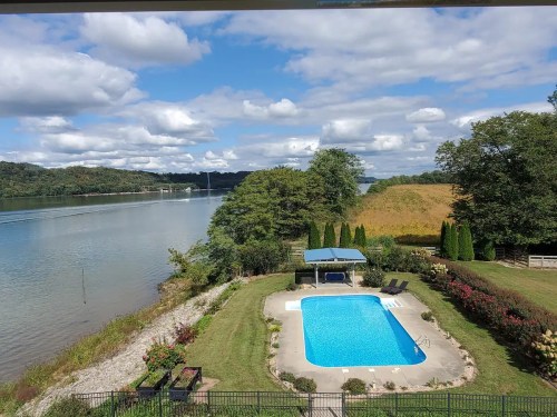 A serene view of a pool by a river, surrounded by greenery and under a partly cloudy sky.