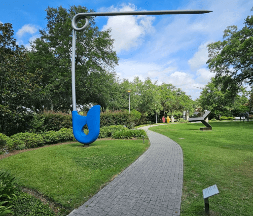 A large blue safety pin sculpture stands beside a winding path in a green park with trees and a cloudy sky.