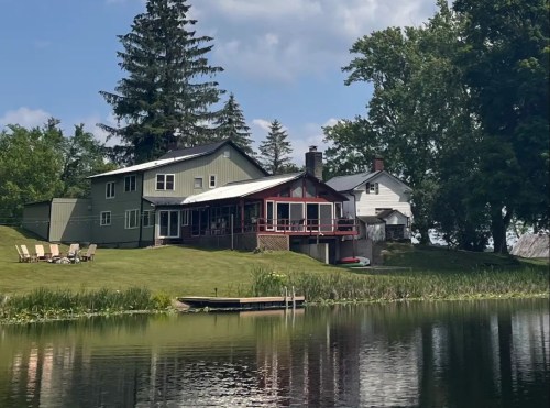 A serene lakeside view of a house with a porch, surrounded by trees and a grassy area, reflecting in the water.