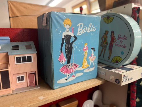 A vintage Barbie lunchbox featuring colorful illustrations of Barbie and friends, displayed on a shelf with toys.