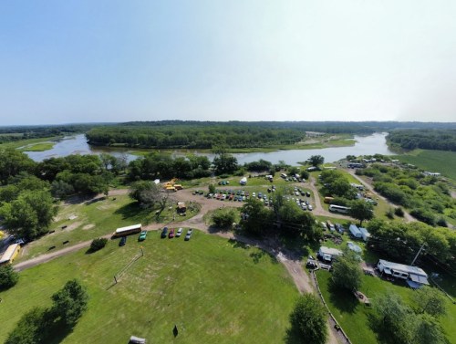 Aerial view of a lush green landscape with a river, surrounded by tents and parked cars in a recreational area.